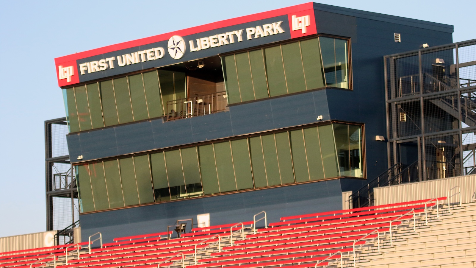 Exterior view of a modern stadium press box with a red signage reading'FIRST UNITED LIBERTY PARK' and blue walls, red seating below