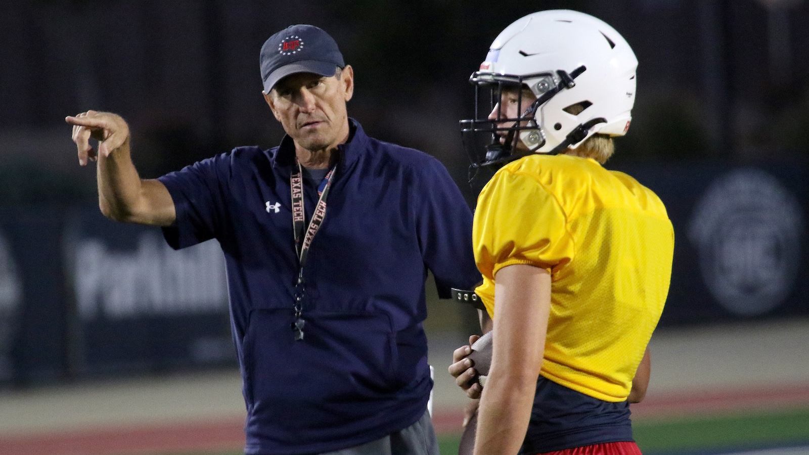 Coach on a football field points and talks to a player in a yellow jersey and helmet, giving directions during practice.