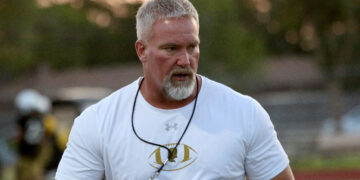 Middle-aged man with gray hair and beard wearing a white athletic shirt, sunglasses on a cord around his neck, outdoors at a sports field.