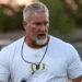 Amarillo High - HC Chad Dunnam 2 | Press Pass Sports Middle-aged man with gray hair and beard wearing a white athletic shirt, sunglasses on a cord around his neck, outdoors at a sports field.