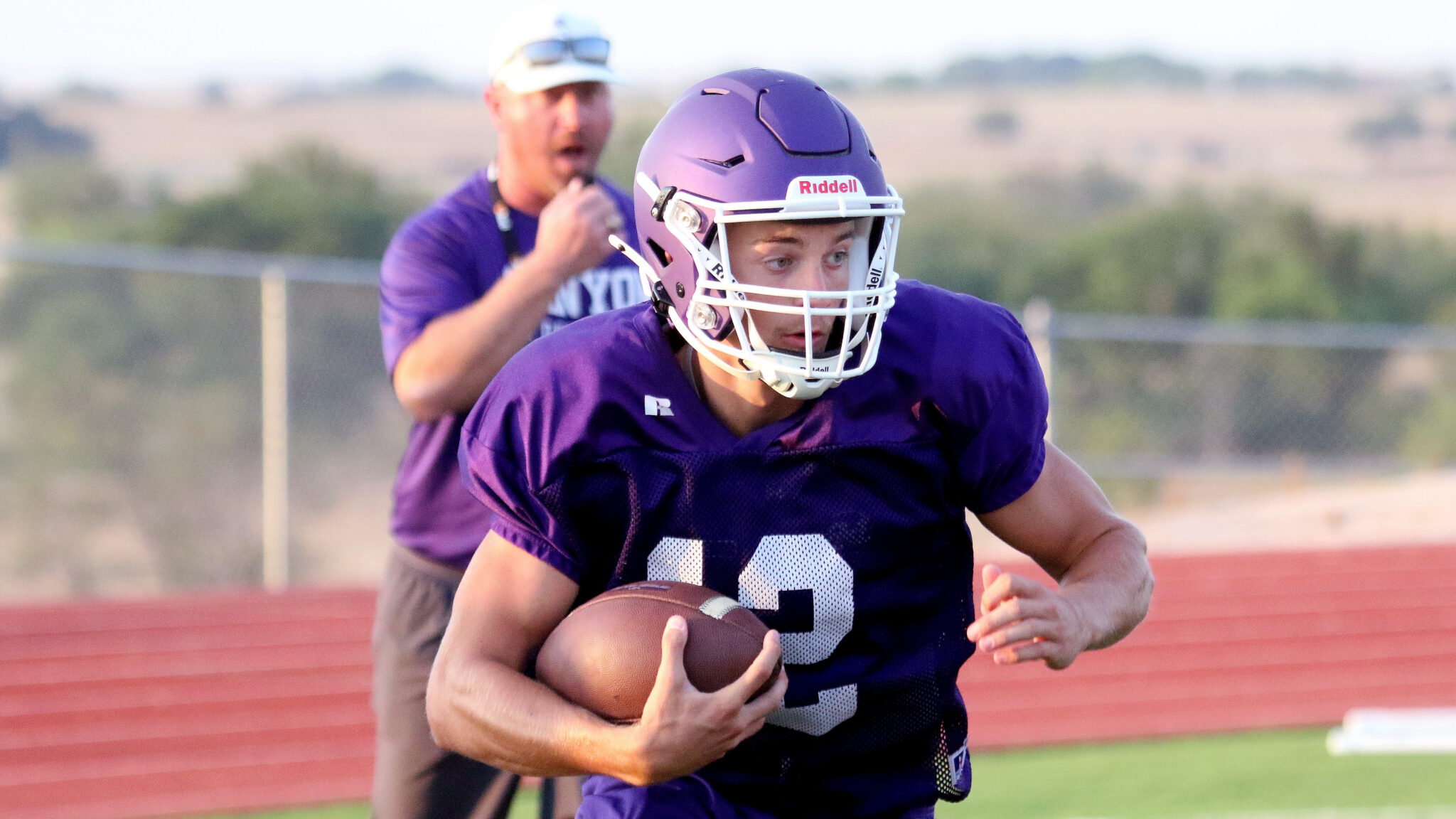 1K6A8921 | Press Pass Sports Football player in a purple jersey carrying a football while running on a track field, helmet on