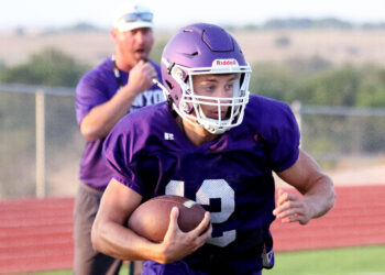 1K6A8921 | Press Pass Sports Football player in a purple jersey carrying a football while running on a track field, helmet on