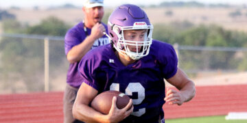 Football player in a purple jersey carrying a football while running on a track field, helmet on