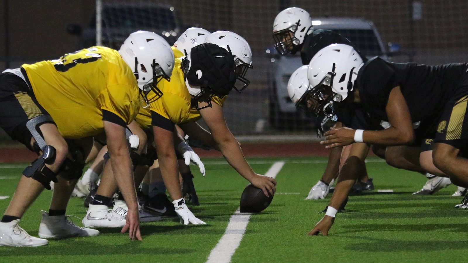 Football players line up at the line of scrimmage during a night game, centers ready to snap the ball between yellow and black teams