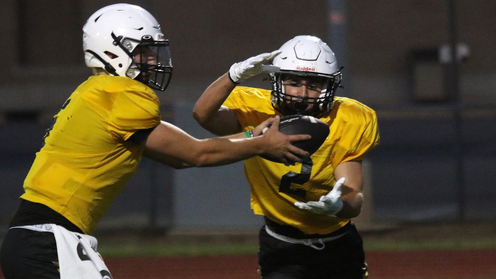 Two football players in yellow jerseys and white helmets reach for a football during a nighttime practice, one guarding the ball.