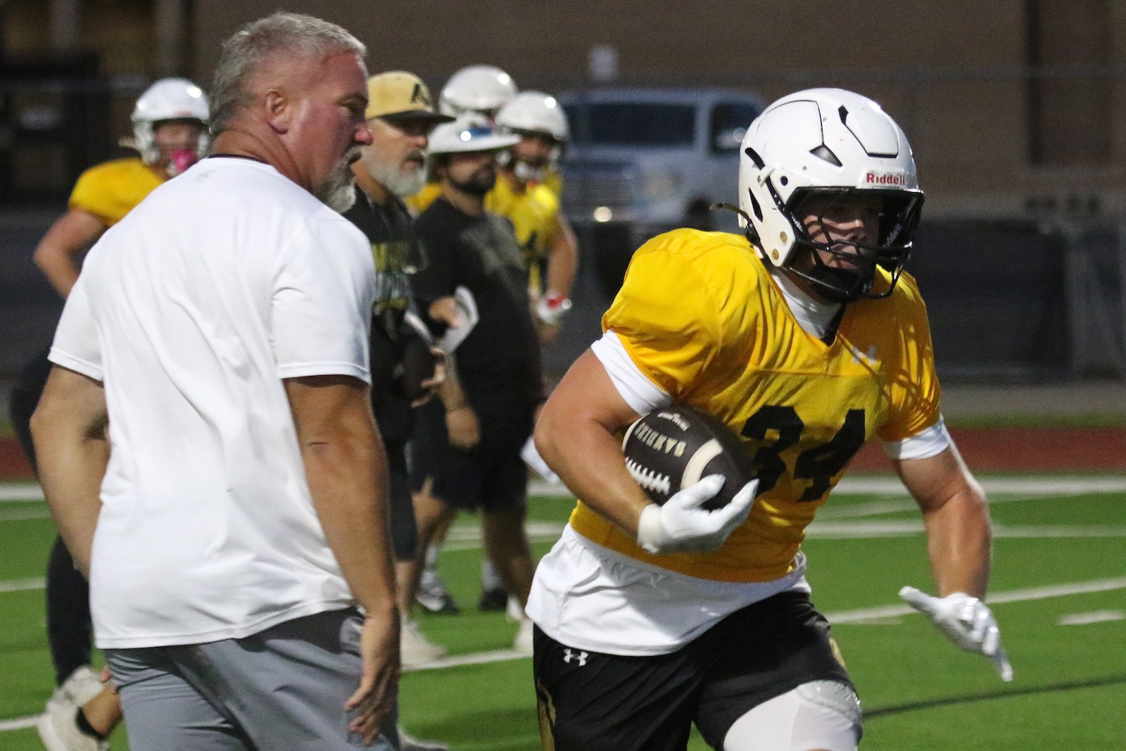 Football player in a yellow jersey and white helmet runs with the ball on a turf field, a coach watching in the foreground as teammates line up in the background.