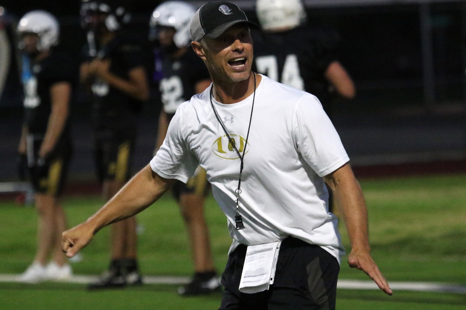 Football coach shouting instructions on a turf field, wearing a white shirt, cap, and whistle with players in the background on night practice.