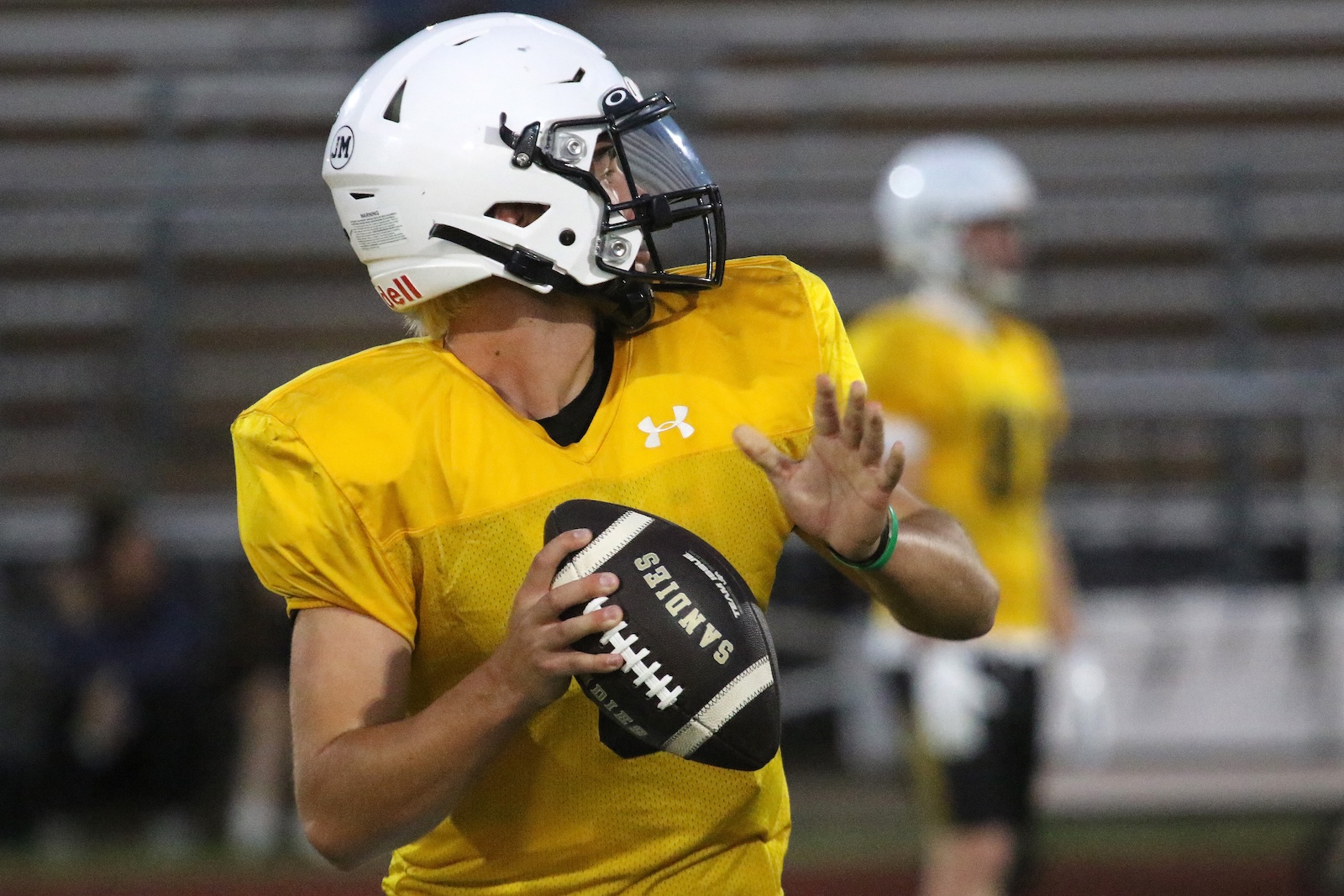 Quarterback in a yellow practice jersey and white helmet prepares to throw a football on a practice field at night.