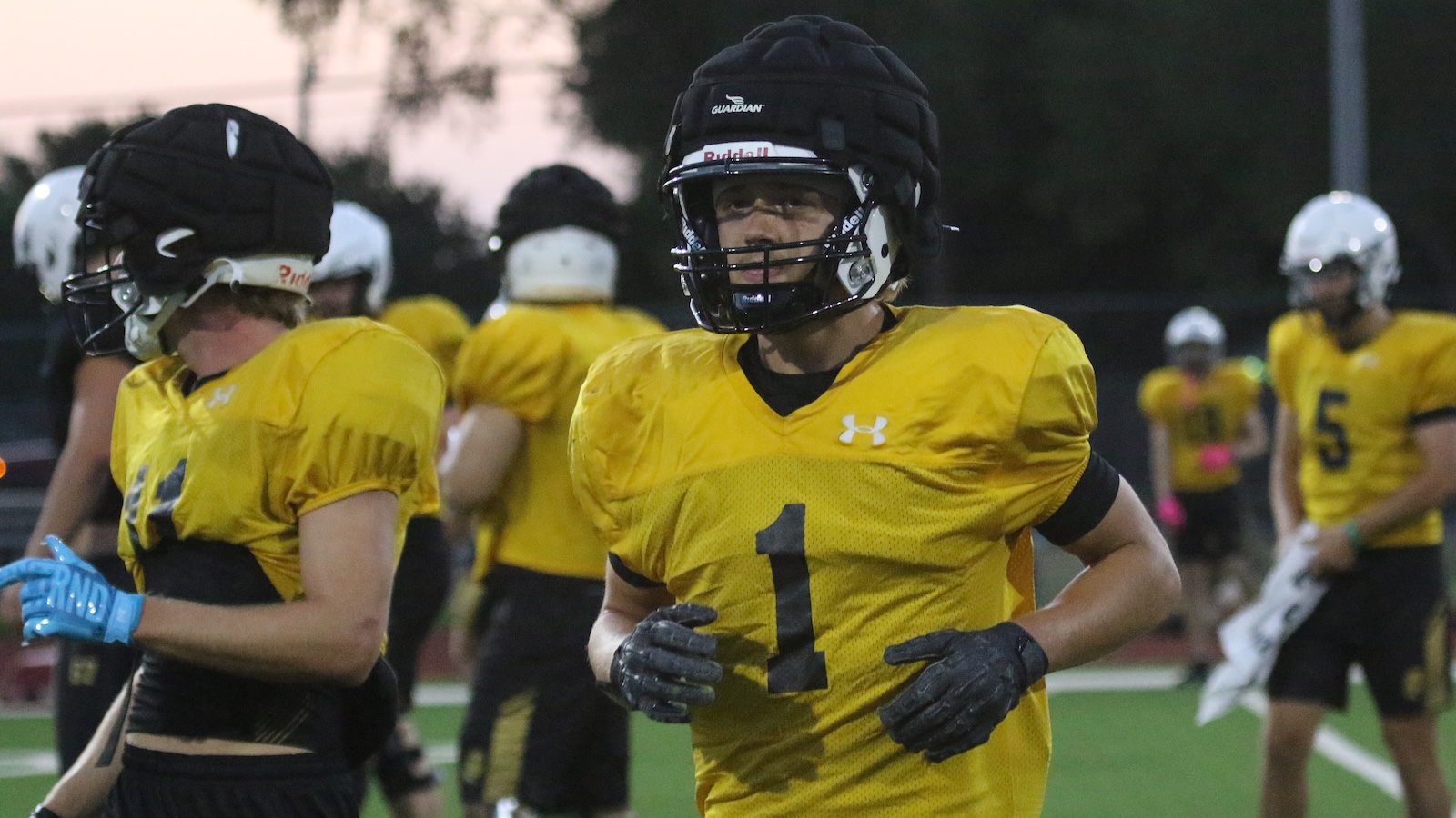 Football players in yellow jerseys and helmets on a field, central player wearing number 1 looks ahead while running.