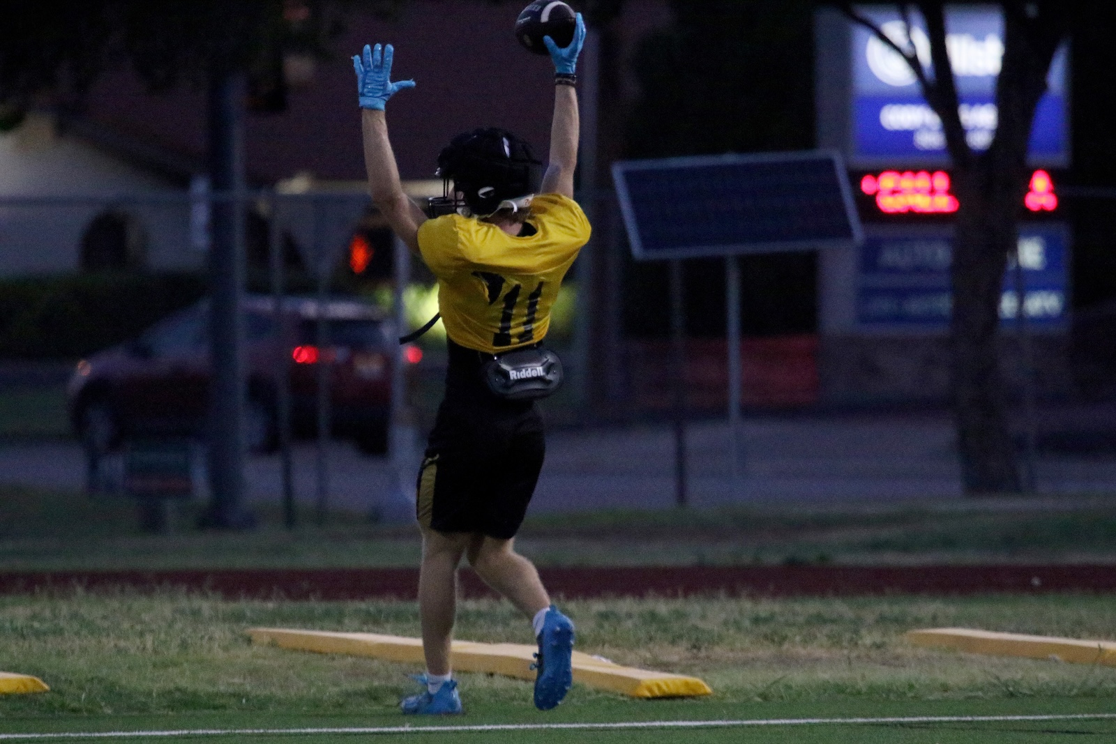 Football player in a yellow jersey (number 11) with blue gloves leaping to catch a ball overhead on a darkened field at dusk.
