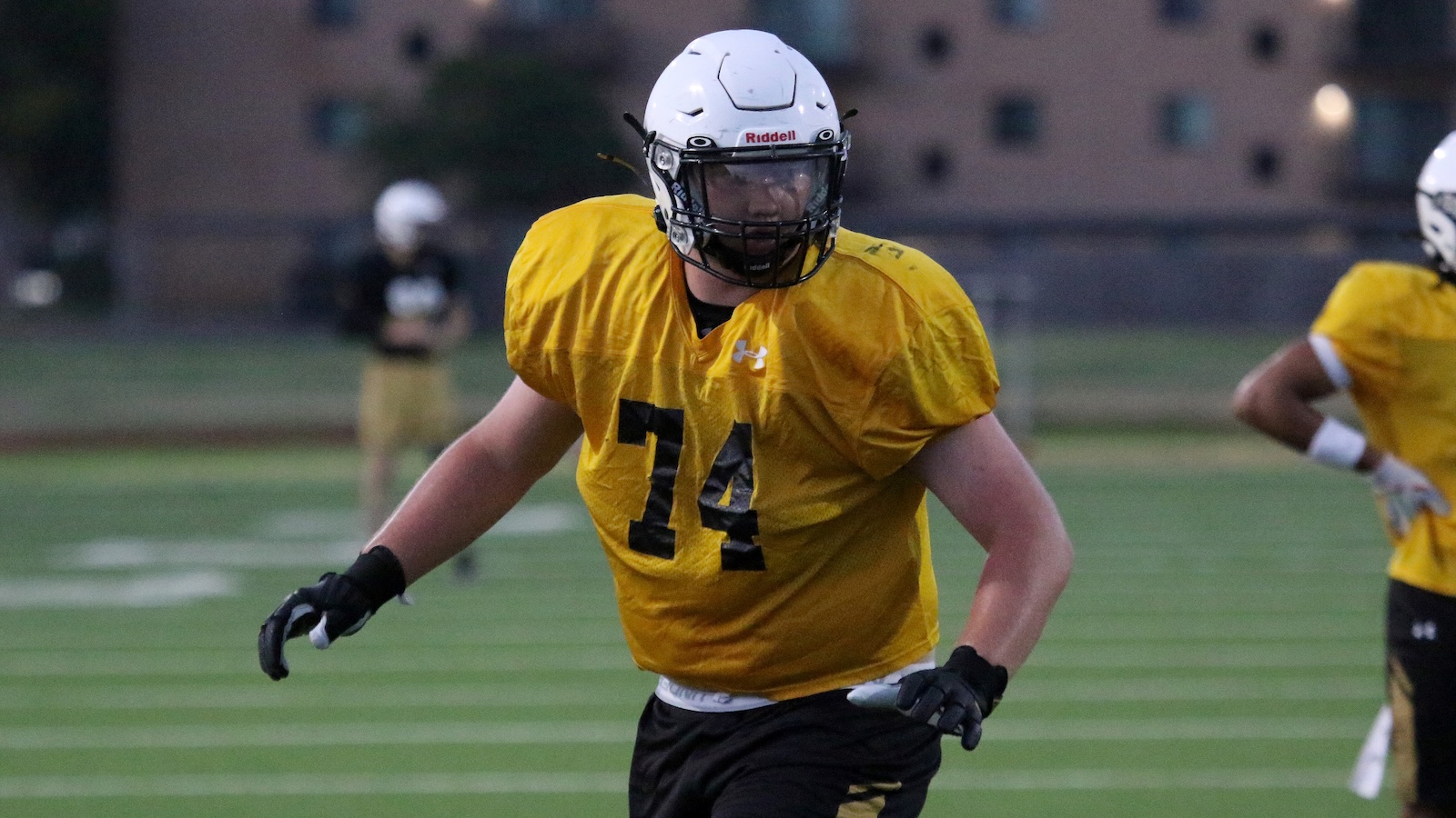 American football player in a yellow jersey number 74 runs on a green field, wearing a white helmet and black gloves during practice.