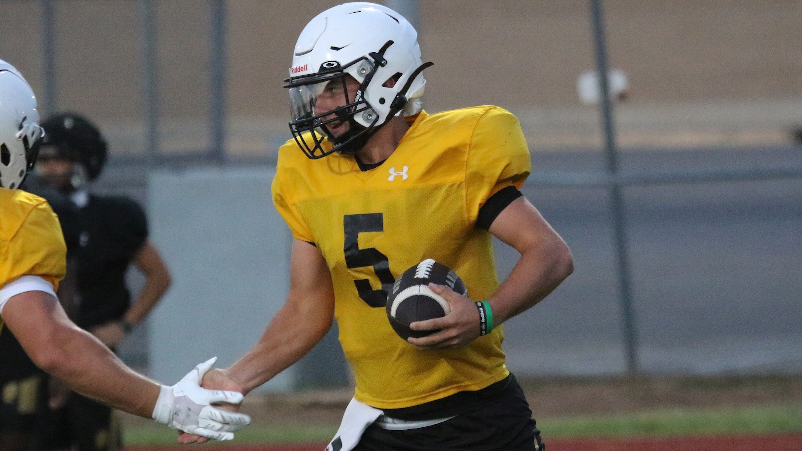 Football player in a yellow jersey with number 5 holding a football during practice, approaching a teammate for a handshake on the field.