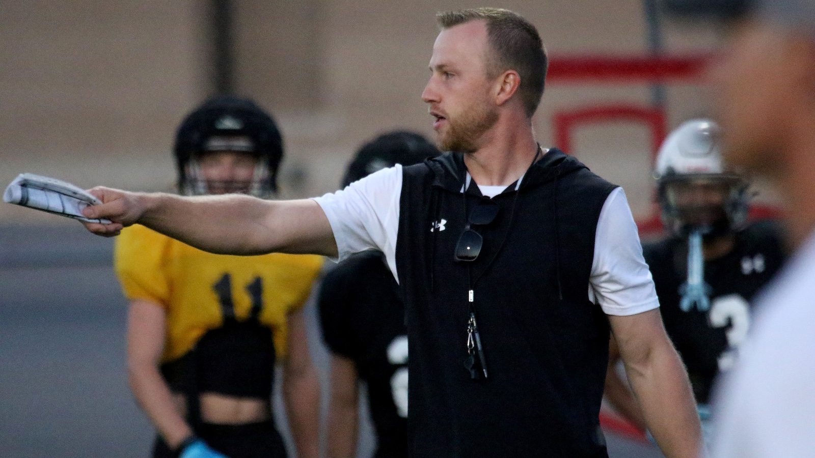 Coach in a black sleeveless hooded shirt points with a clipboard in hand on a football field during practice.