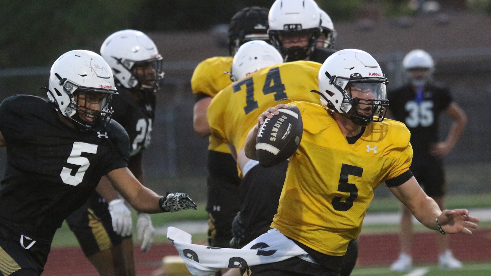 Football player in a yellow jersey #5 runs with ball, pursued by players in black jerseys during a game.