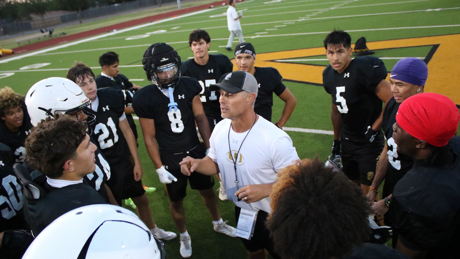 Football coach giving a pep talk to a huddled team on the green turf field during practice.