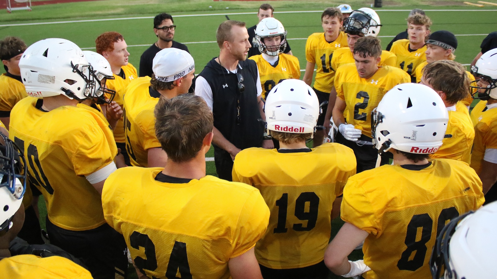 Youth football team in yellow jerseys form a huddle around their coach on the field, listening intently during a timeout or pep talk.