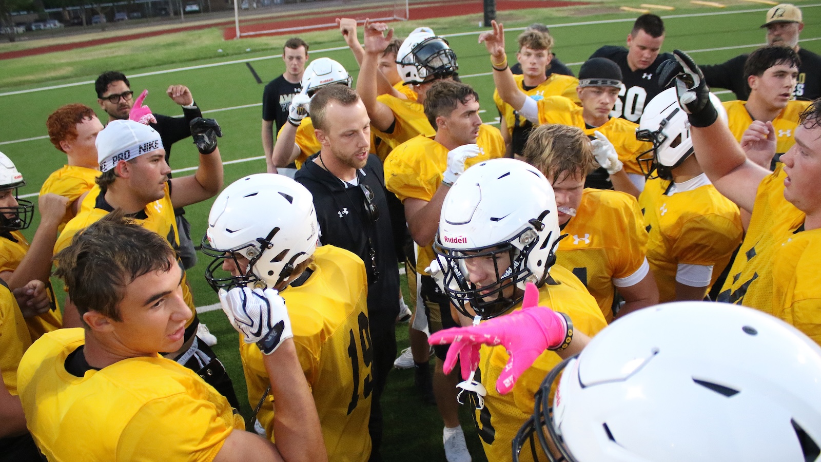 Football team in yellow uniforms huddles on a grassy field, players wearing white helmets as coach gives instructions.