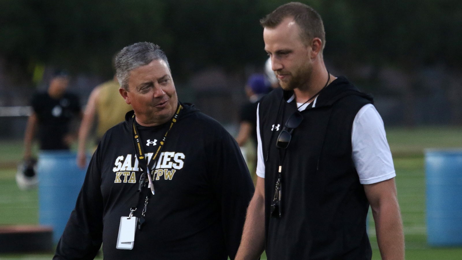 Two men wearing black team gear walk and chat on a grassy sports field, lanyards around their necks, evening light. They appear to be coaches or staff.