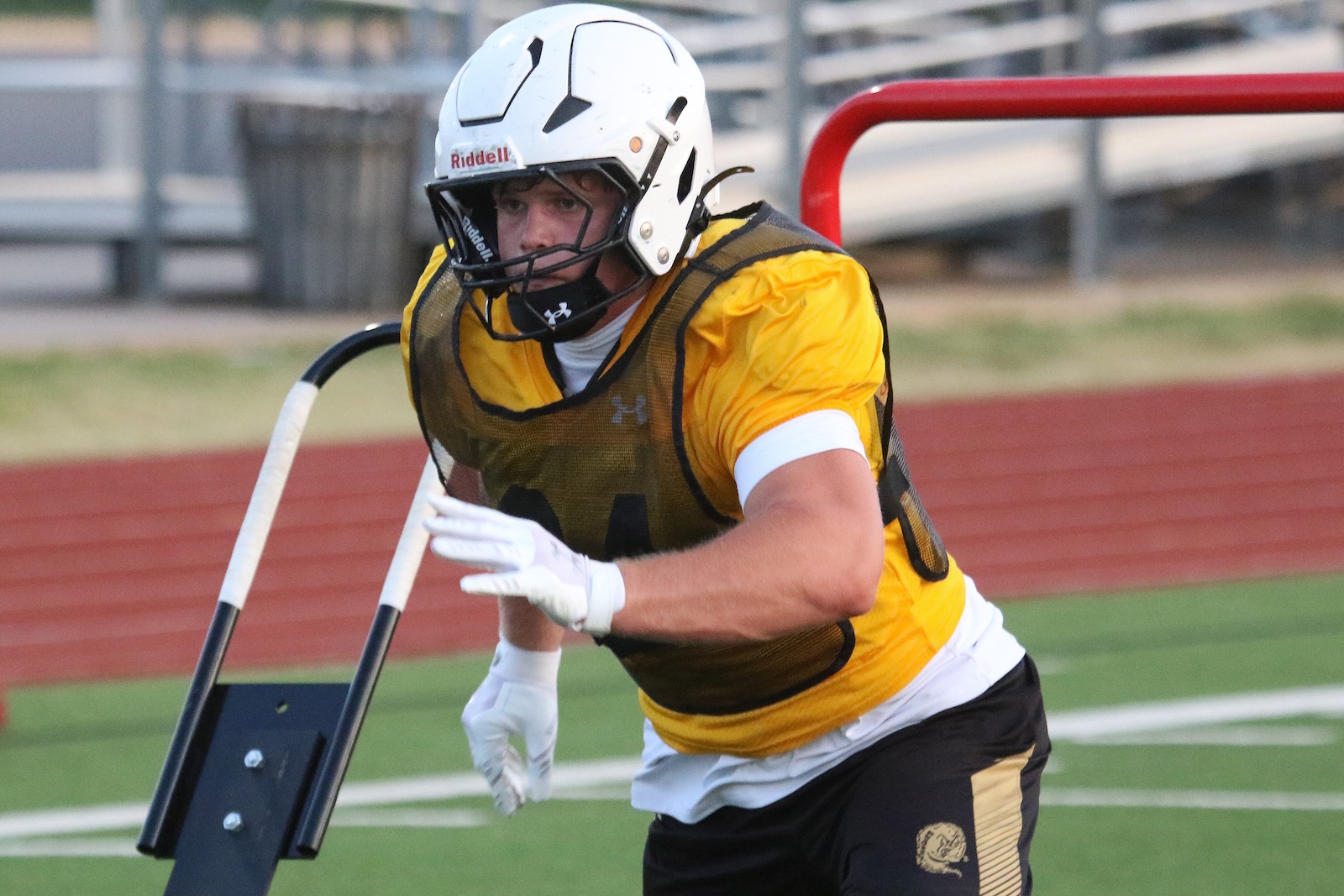 Football practice: player in a yellow jersey and helmet running with a blocking shield attached to a metal frame on a track field.