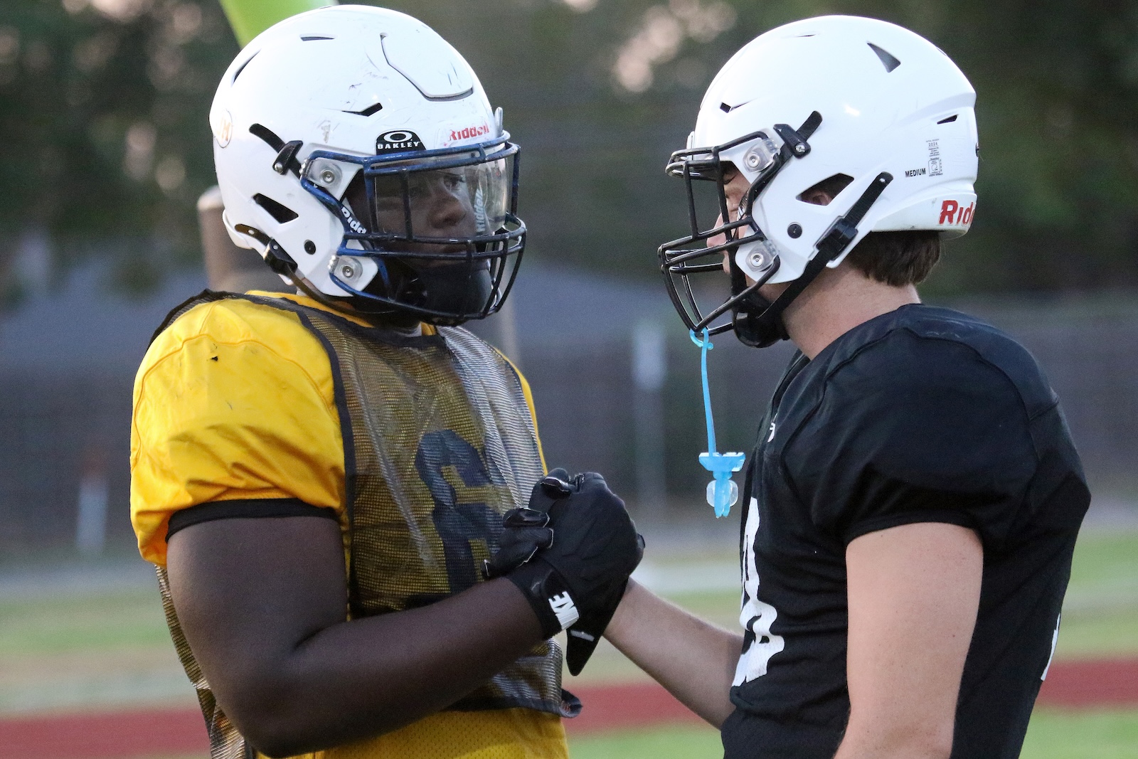 Two football players wearing helmets face each other and touch fists on a field, one in yellow practice gear and the other in black.