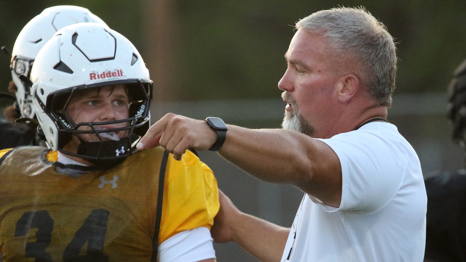 Football coach gives instructions to a football player in an orange practice jersey and white helmet during drills on the field.