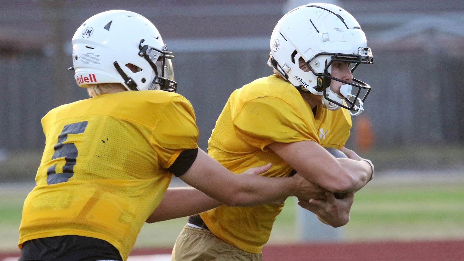 Two football players in yellow practice jerseys and white helmets engage in a drill, one grabbing the other from behind during a tackle sequence.
