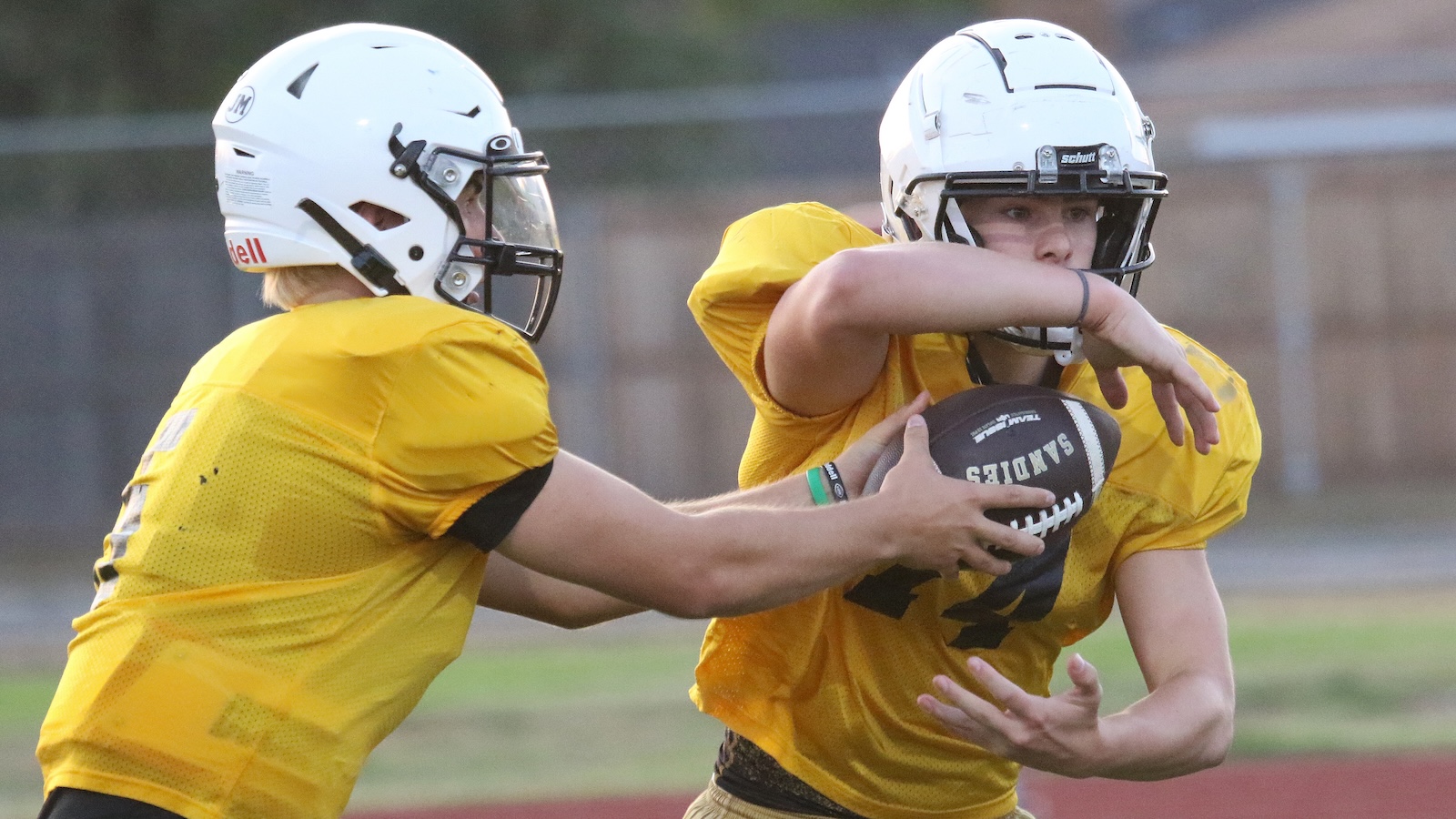 Two football players in yellow practice jerseys fight for control of a ball during a drill on a field, both wearing white helmets.
