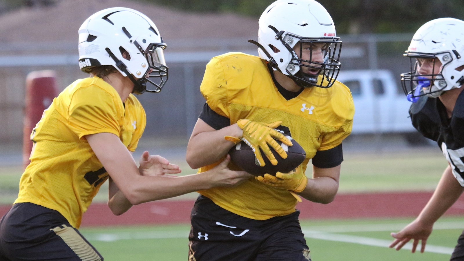 Three football players in yellow jerseys and white helmets engage in a practice drill on a field; the center player cradles a football while another player reaches to block him.