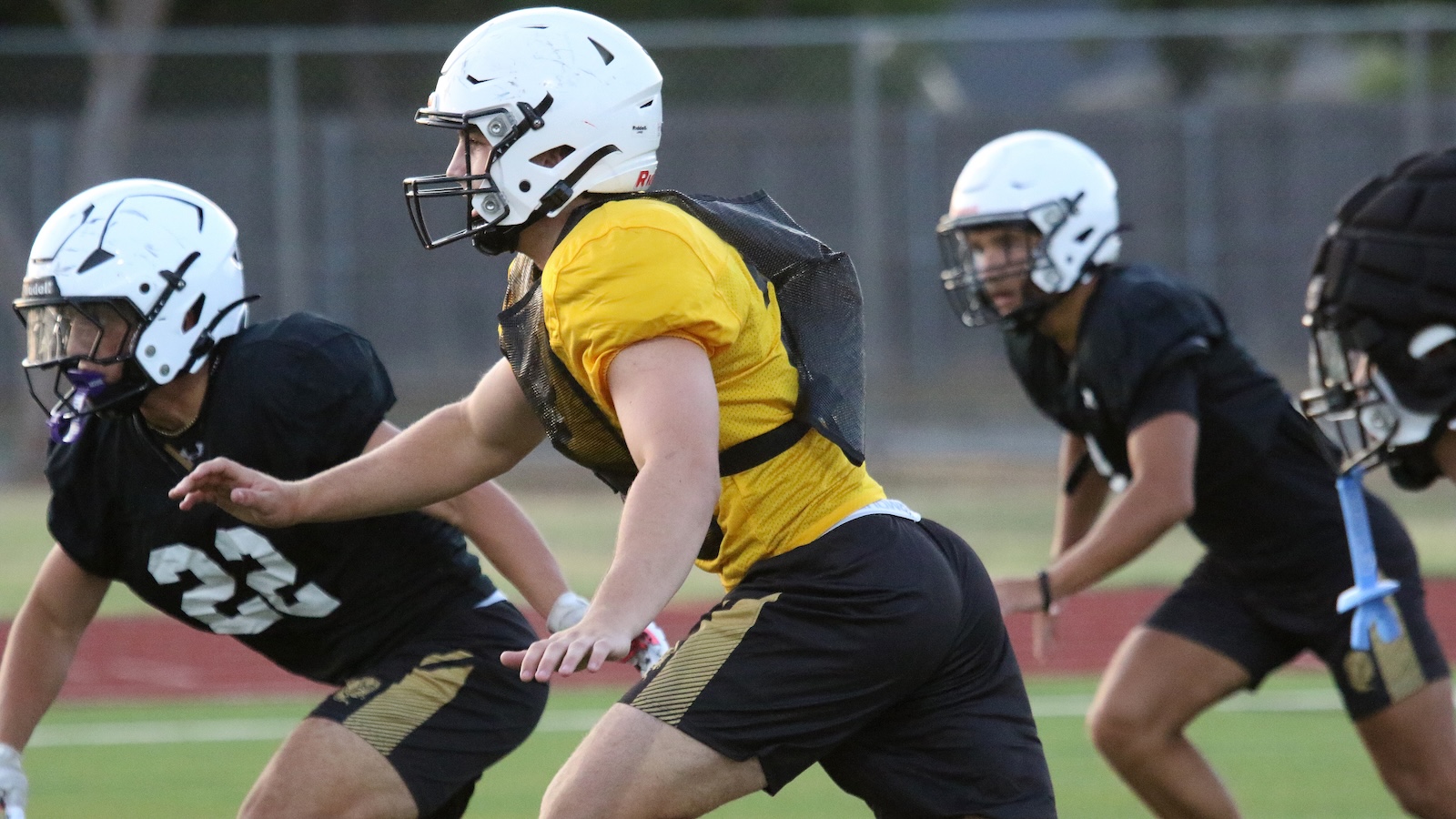 Youth football players in helmets and pads sprinting on a practice field; central player wears a yellow jersey and black shorts with a mesh vest catching the action