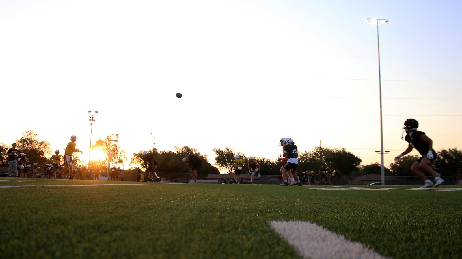 Youth football players practice on a green turf field at sunset, with a football in midair.