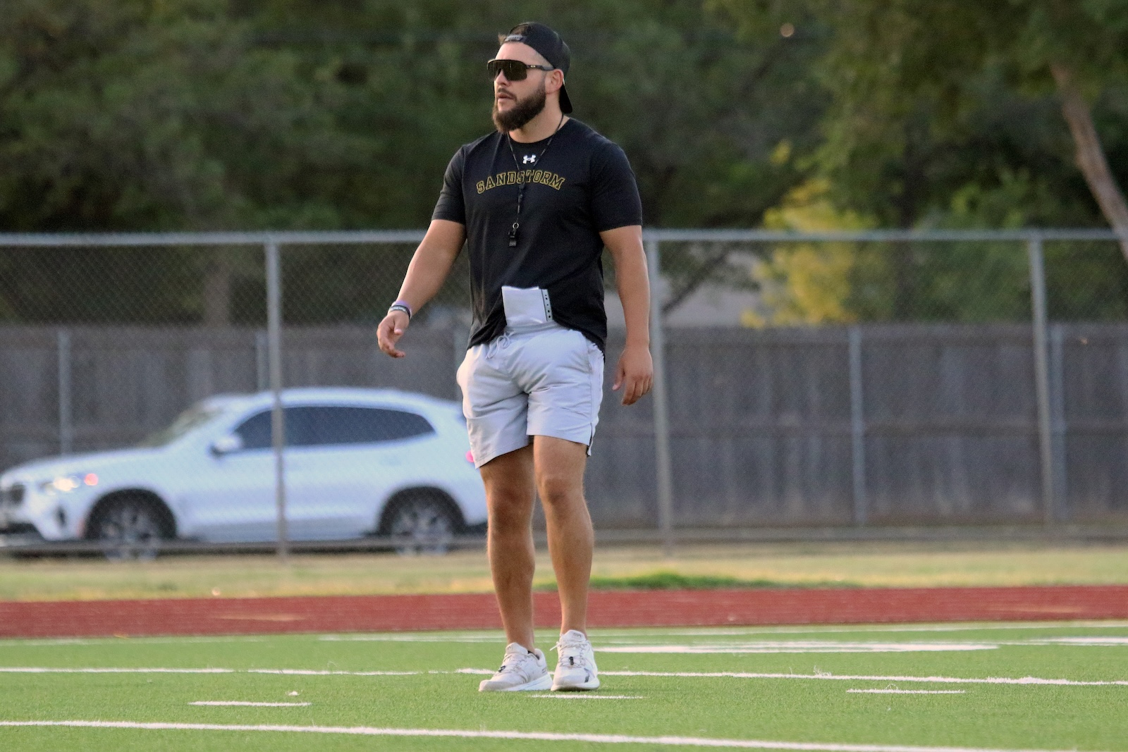 Man wearing a black t‑shirt, cap backward and sunglasses stands on a football/soccer field with a notebook tucked into his waistband.