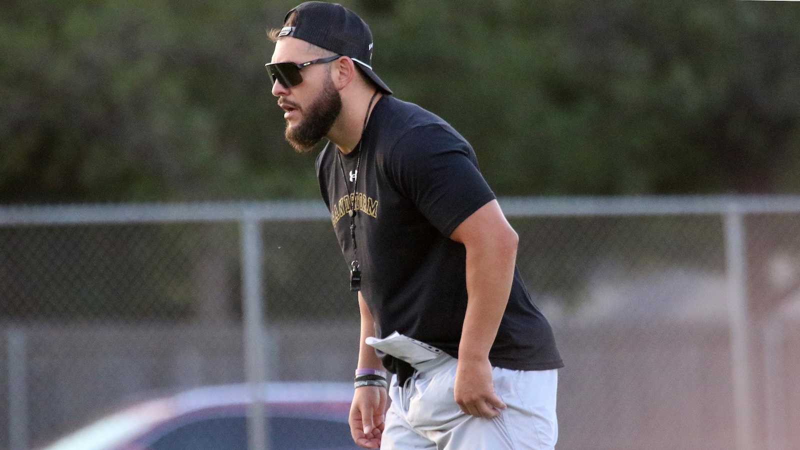 Athletic coach wearing a black t-shirt, backwards cap, sunglasses, and a whistle, bending slightly on a field with a chain-link fence behind him.