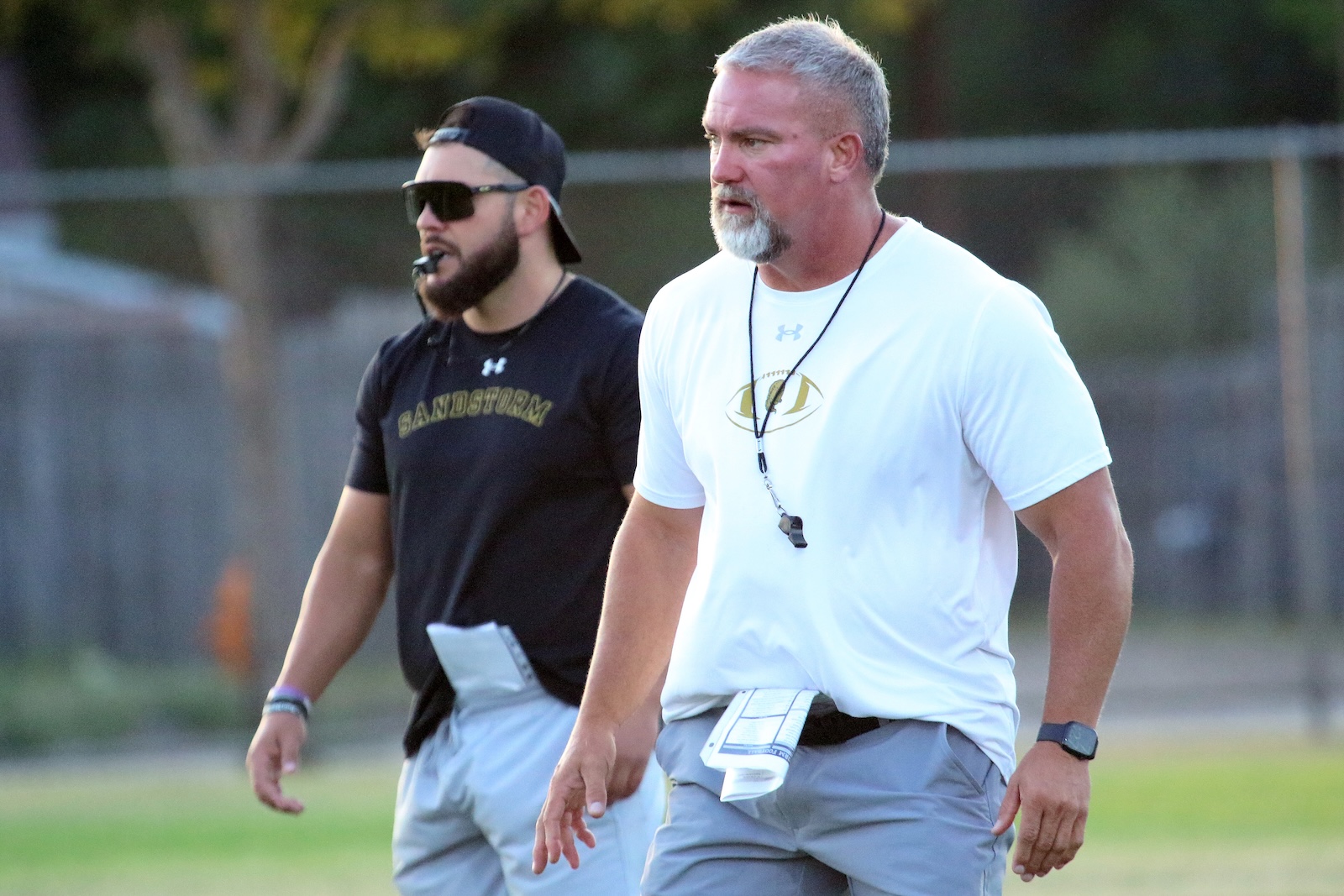 Two adult men walking on a sports field; one in a white shirt with a whistle, the other in a black shirt and cap.