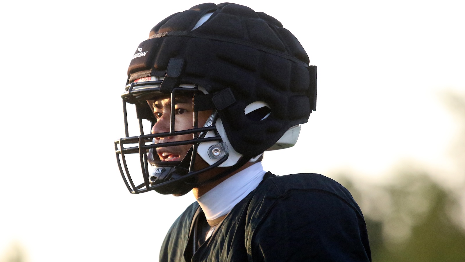 American football player wearing a black protective helmet with facemask, looking to the left on a sunlit field