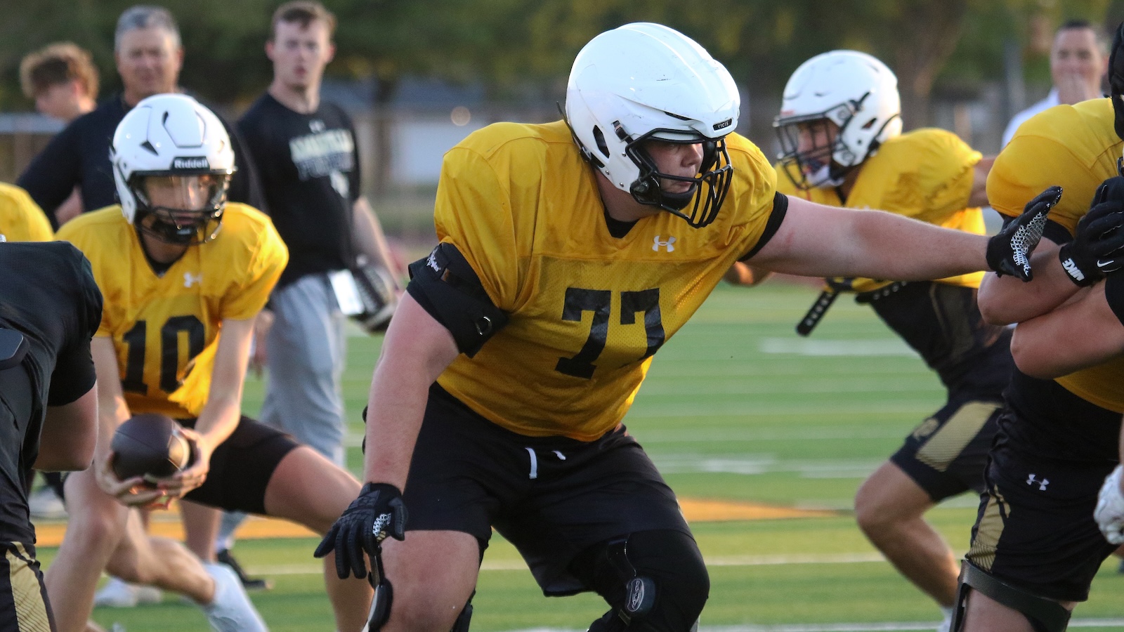 American football player in a yellow jersey (#74) lunges forward during a drill on a green field with teammates in the background.