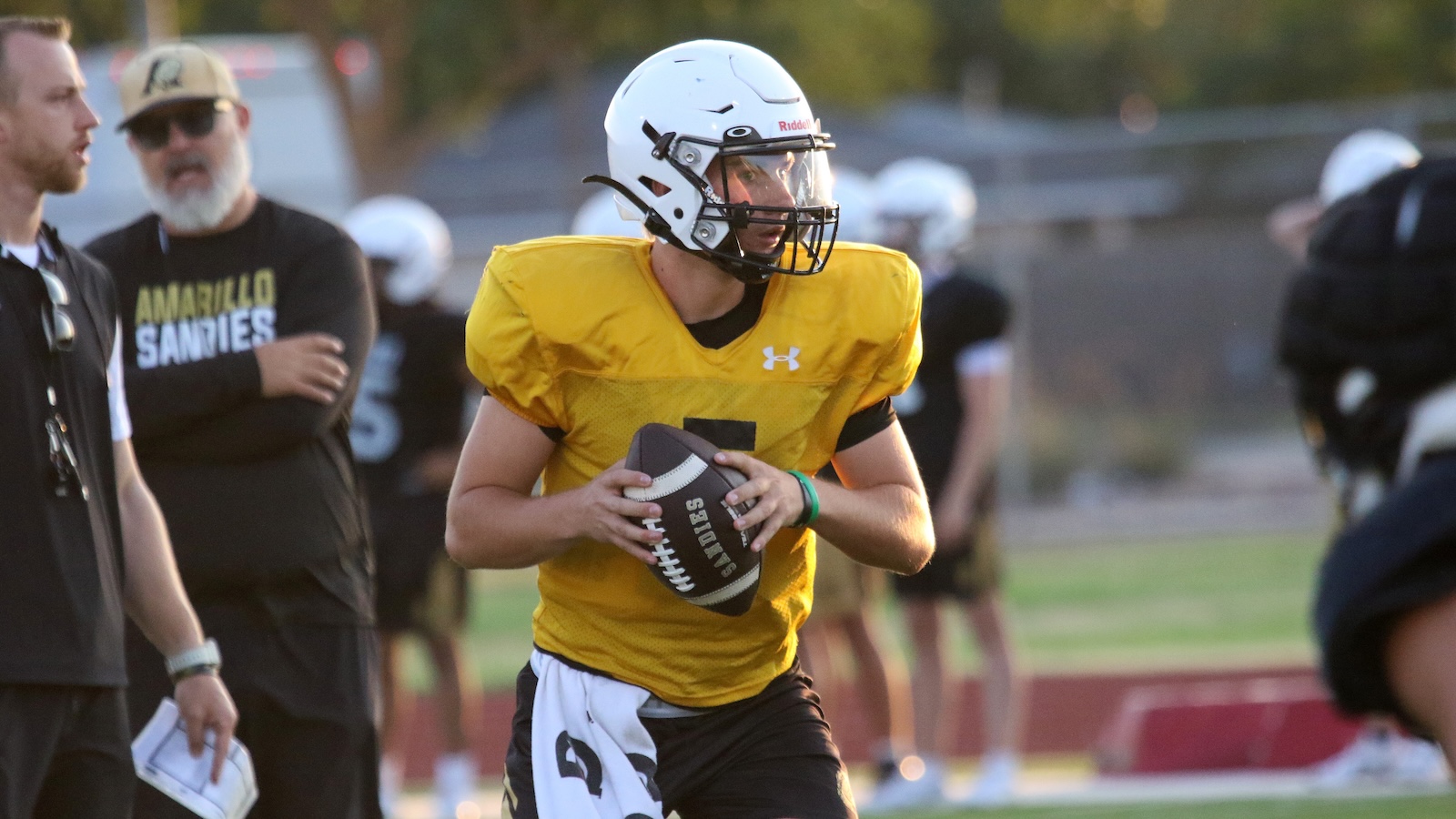 Football player in a yellow practice jersey and white helmet holding a football, mid-run on the field with teammates in the background.] ,...