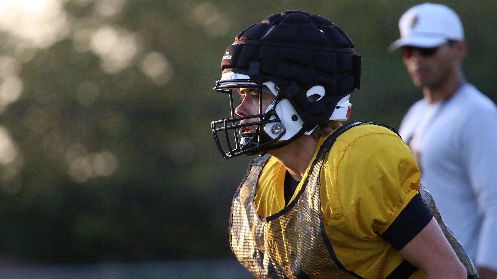 Football player in a yellow practice jersey with a protective mesh vest, helmet on, ready for drills on the field; a coach is blurred in the background.