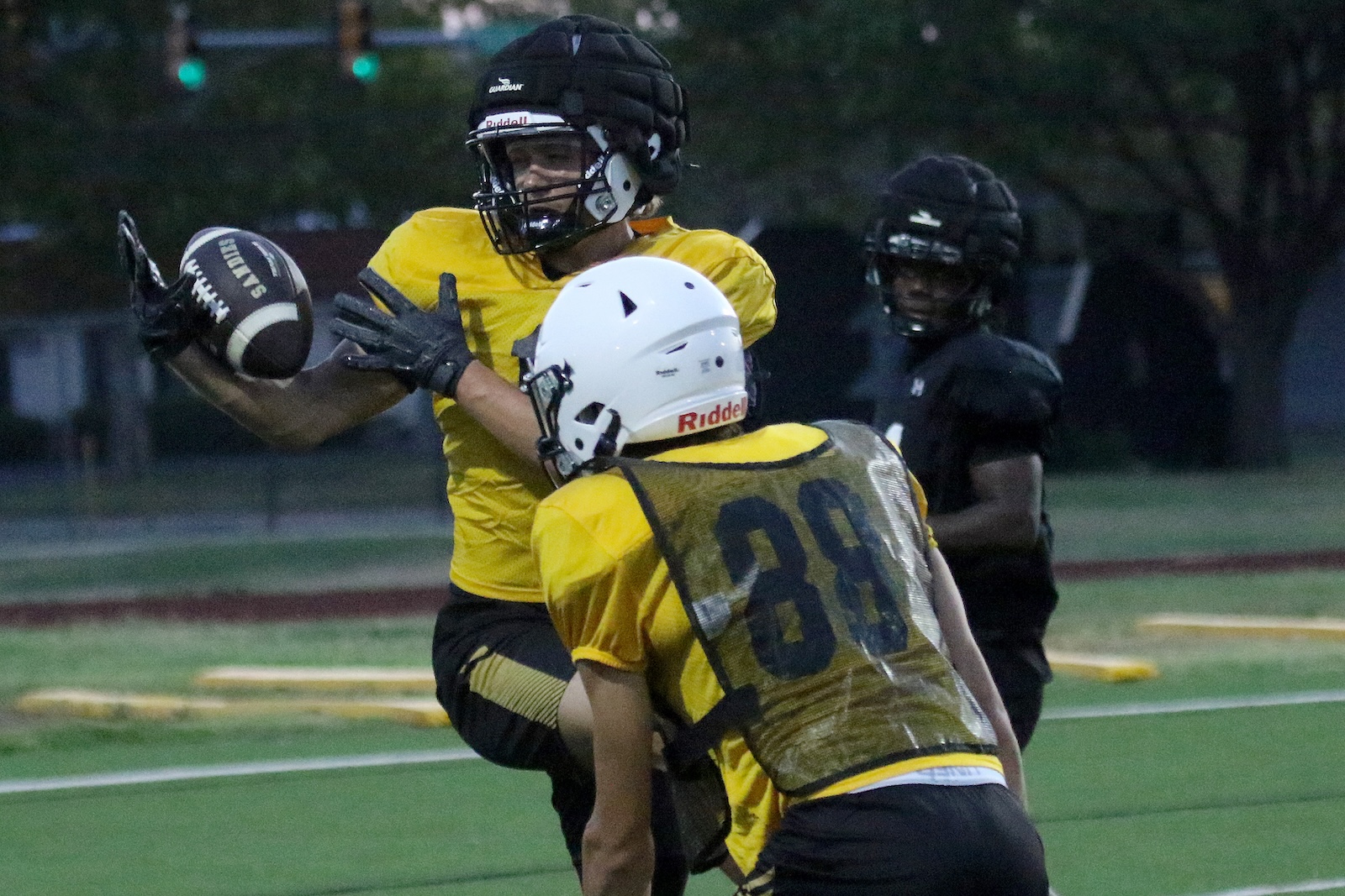 Two youth football players in yellow jerseys contest a play on a turf field at dusk; one reaches to catch a black football while a teammate in a white helmet blocks nearby.