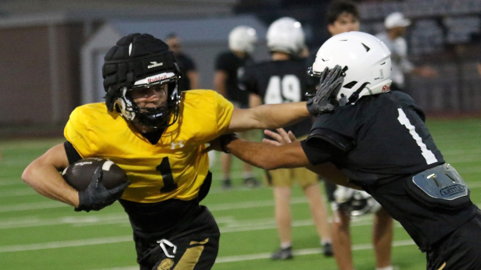 Football player in a yellow jersey runs with the ball while a defender in black reaches to tackle him on a green field.