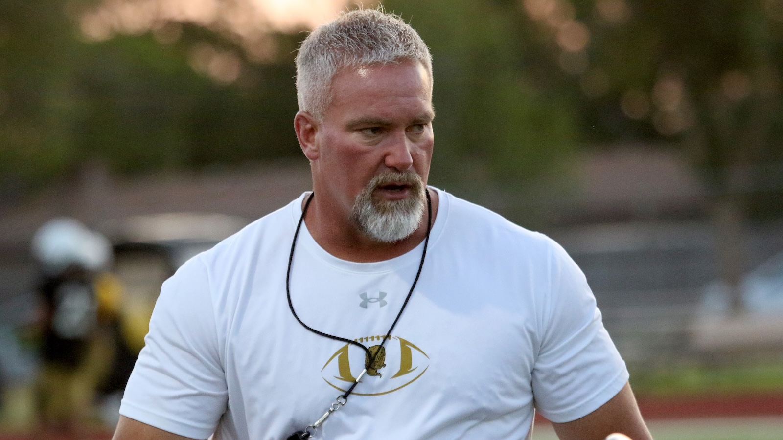 Mid‑aged man with gray hair and a gray beard wearing a white athletic T‑shirt on an outdoor sports field, looking to the side.