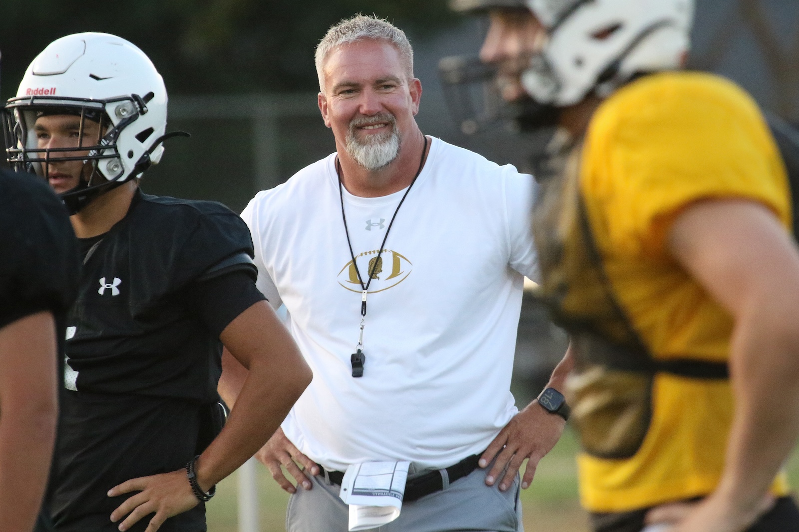 Coaching scene: a smiling football coach with a whistle around his neck stands with players in helmets during practice on a field.