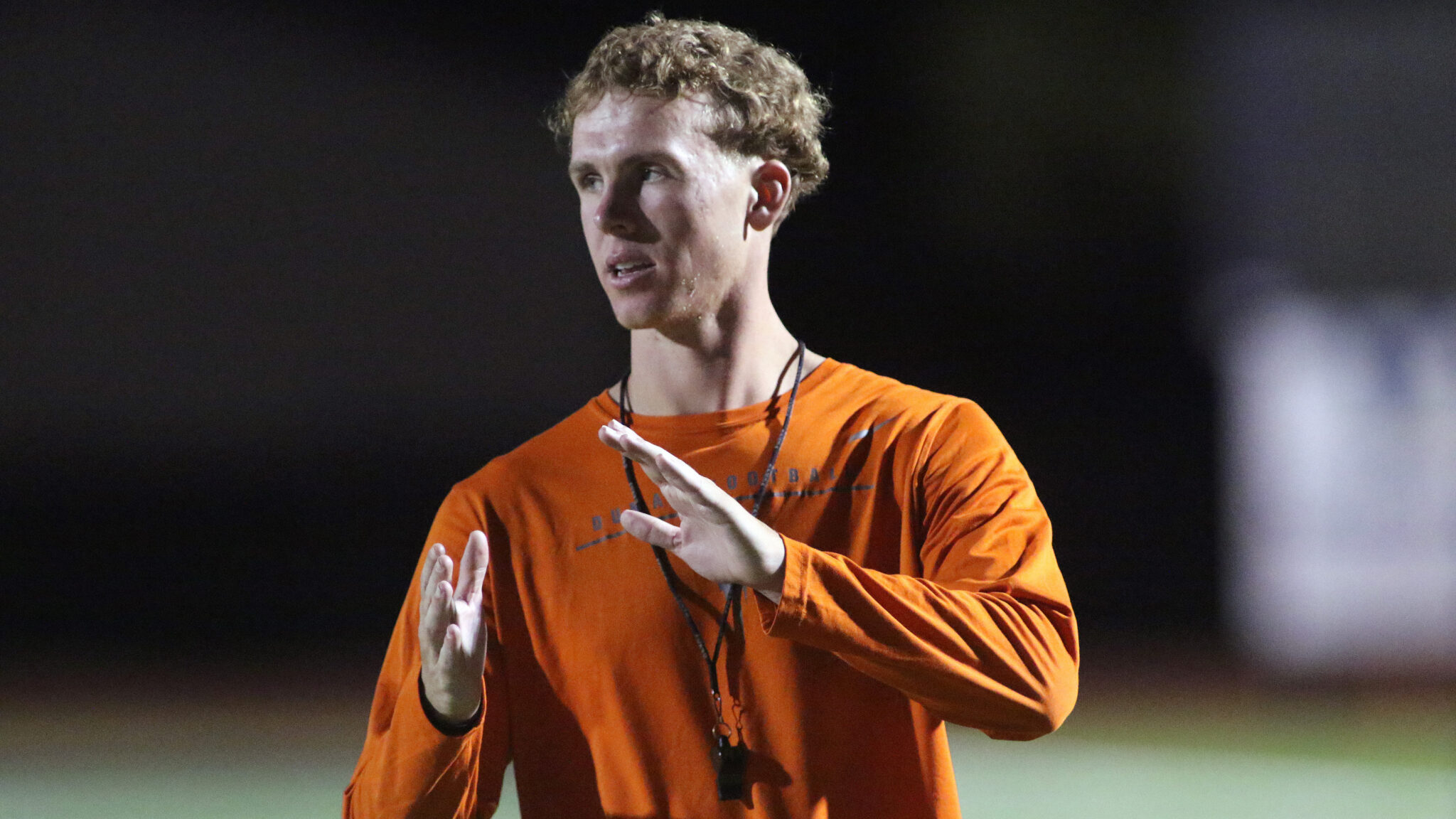 Dumas - Grow Your Own - Hayden Waddell | Press Pass Sports Person in an orange long-sleeve shirt speaks or gestures with hands on a dark, outdoor field at night.