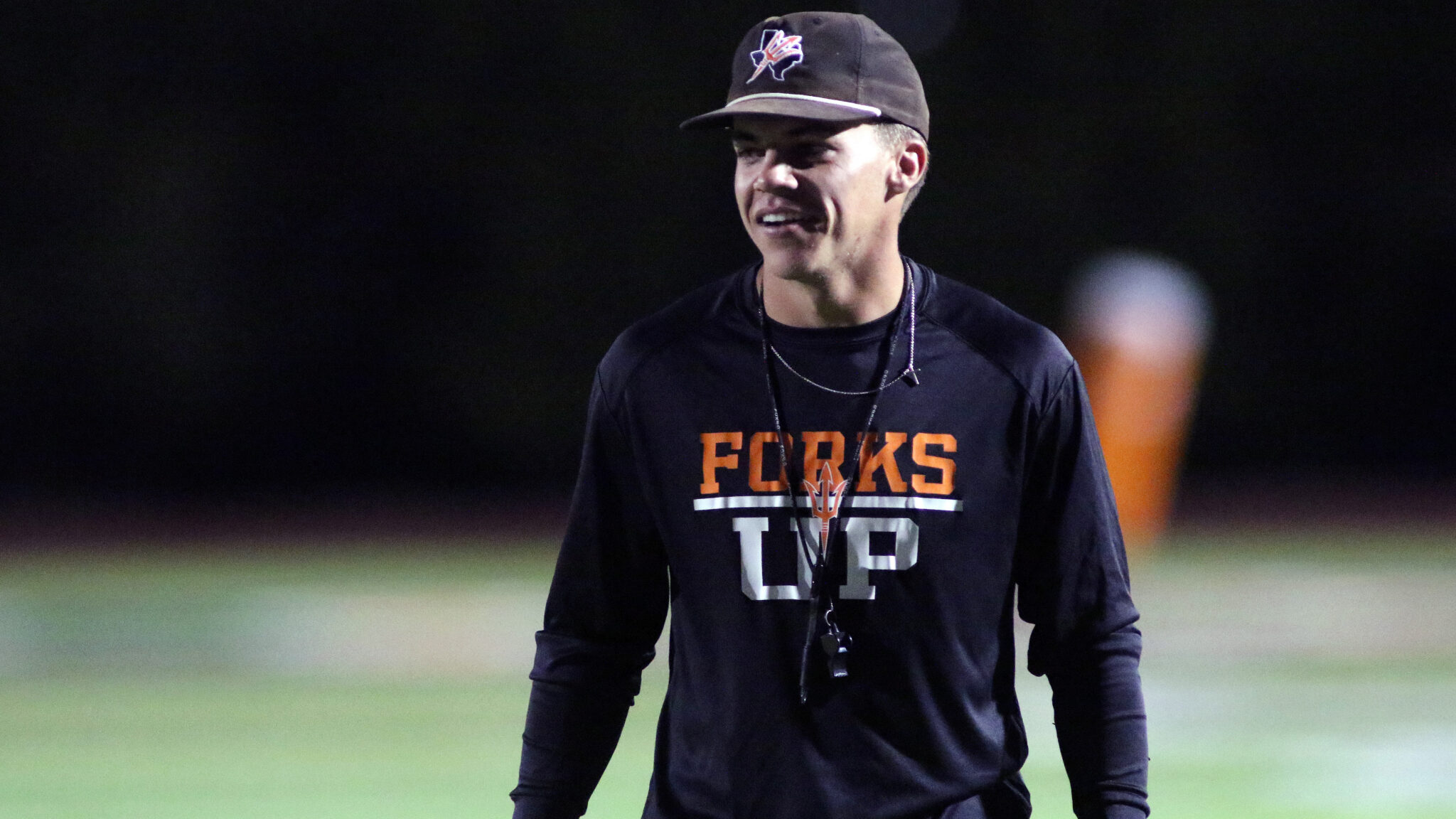 Dumas - Grow Your Own - Parker Dunnam | Press Pass Sports Man wearing a black long-sleeve shirt with orange'FORKS' lettering stands on a dimly lit sports field at night, smiling slightly. Cap visible on head.
