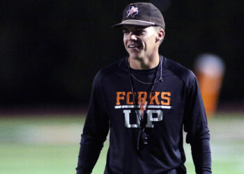 Dumas - Grow Your Own - Parker Dunnam | Press Pass Sports Man wearing a black long-sleeve shirt with orange 'FORKS' lettering stands on a dimly lit sports field at night, smiling slightly. Cap visible on head.