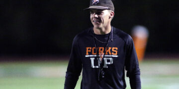 Man wearing a black long-sleeve shirt with orange 'FORKS' lettering stands on a dimly lit sports field at night, smiling slightly. Cap visible on head.