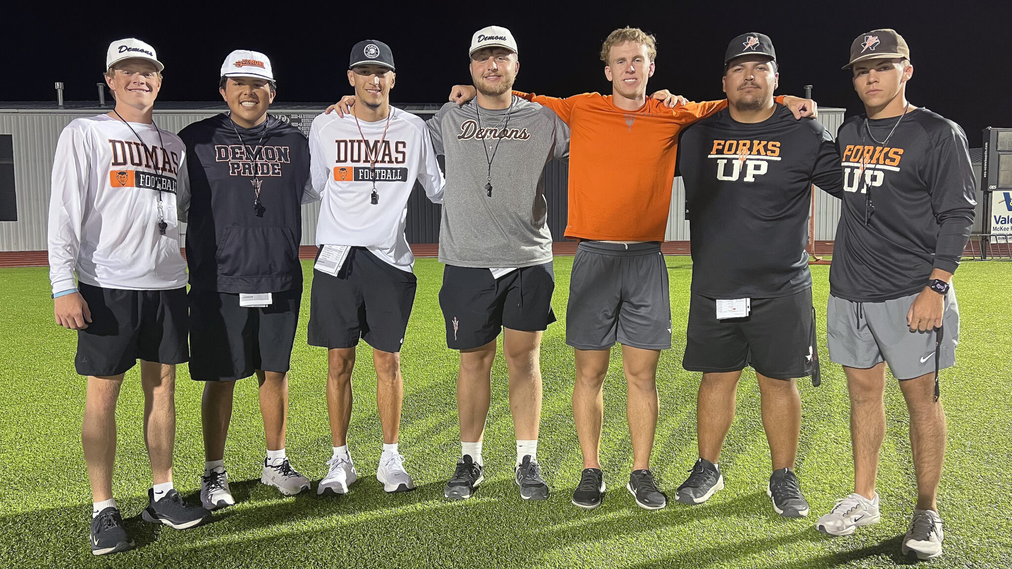 Dumas - Grow Your Own | Press Pass Sports Seven young men in football gear stand with arms around each other's shoulders on a well-lit field at night, posing for a group photo.