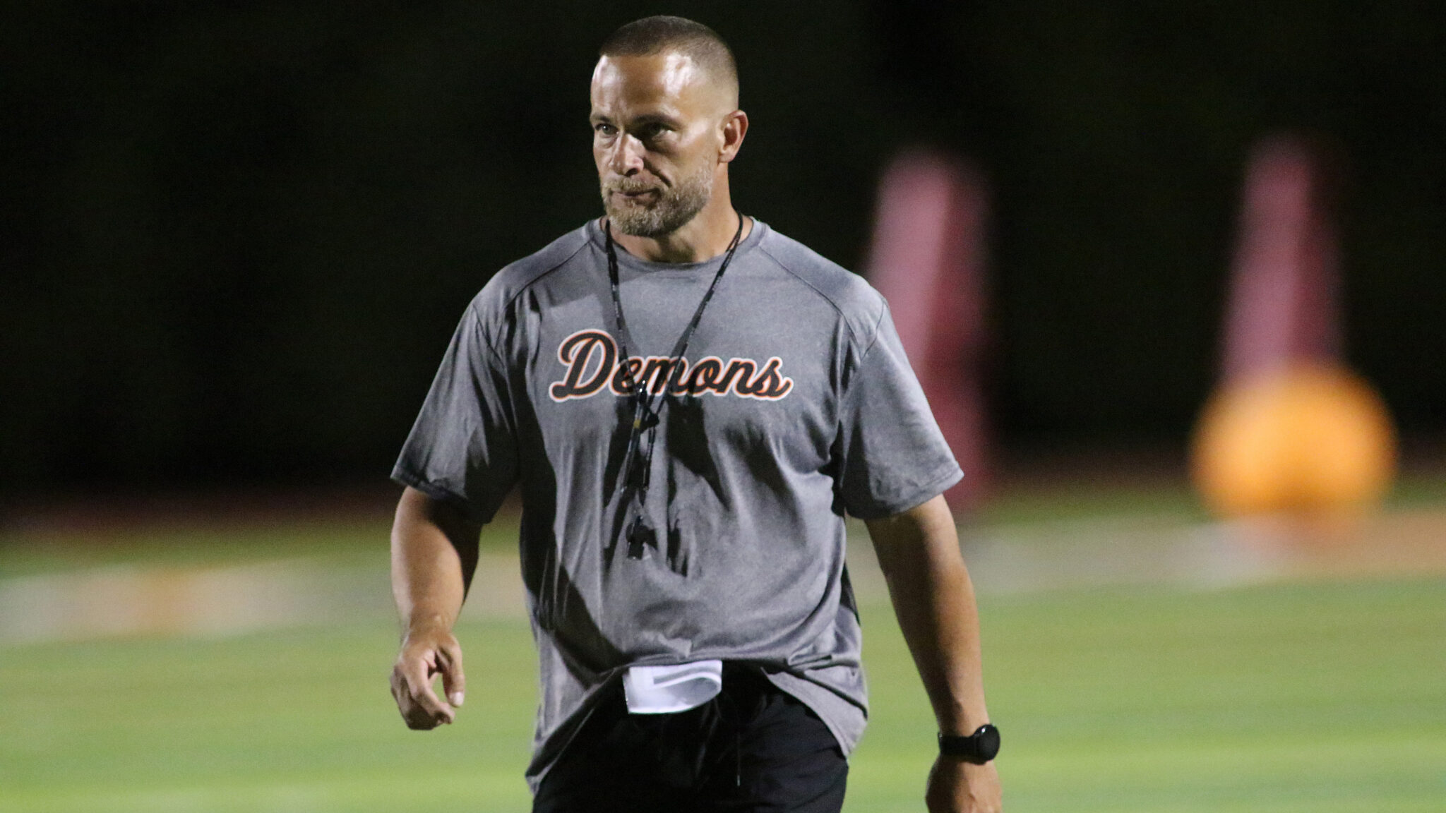 Dumas - HC Aaron Dunnam 2 1 | Press Pass Sports Adult man in a gray Demons shirt walking on a lit sports field at night, wearing a whistle lanyard around his neck.