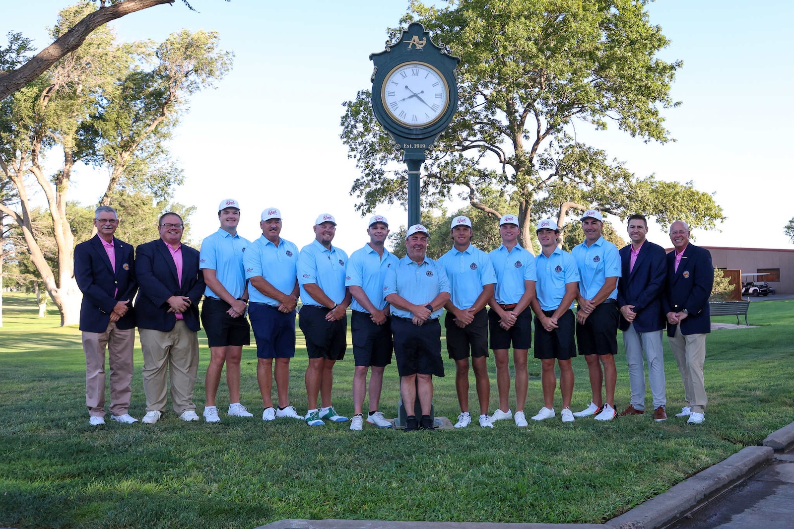 Golf team posed outdoors in matching light-blue polo shirts and caps, flanked by two men in navy blazers, standing in front of a large clock tower in a park.