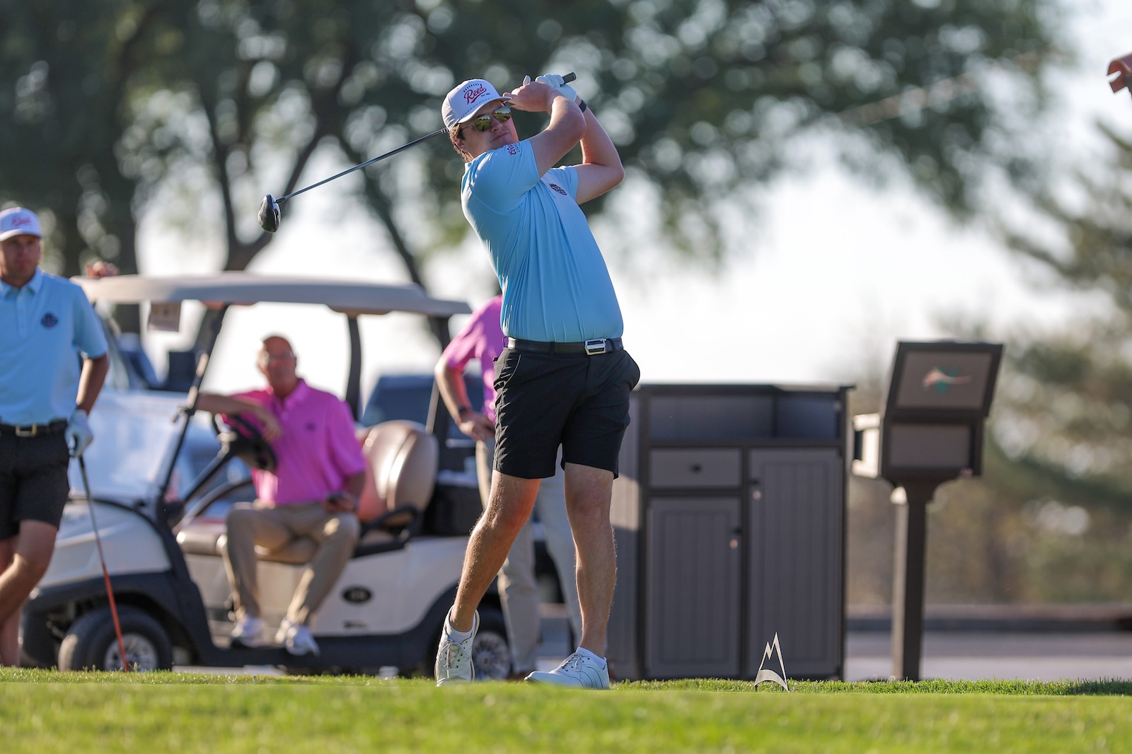 Golfer in a blue polo mid-swing on a sunny golf course, with a golf cart and teammates in the background behind.