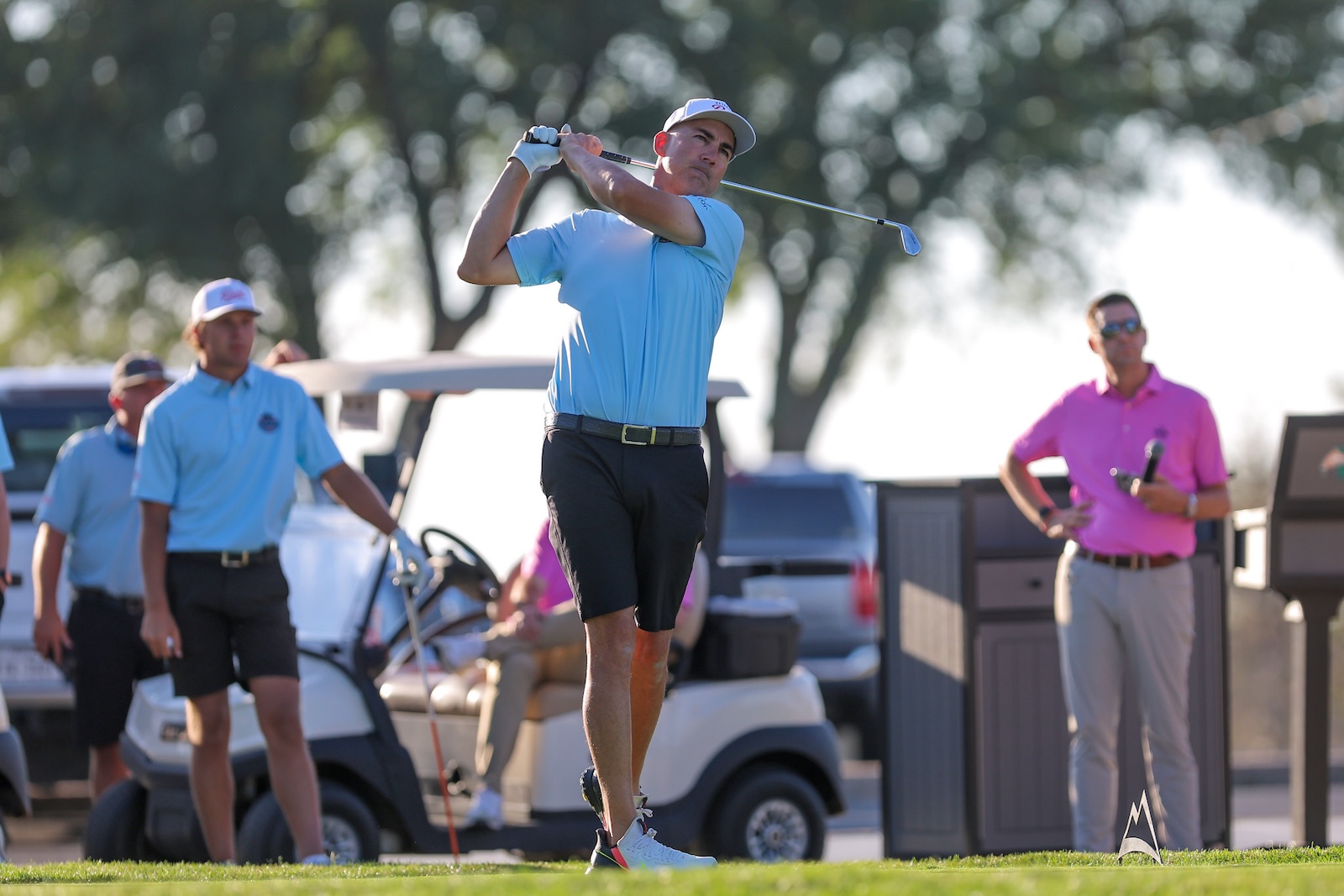 Golfer in a light blue polo and white cap mid-swing on a sunny golf course, with spectators and golf carts in the background.